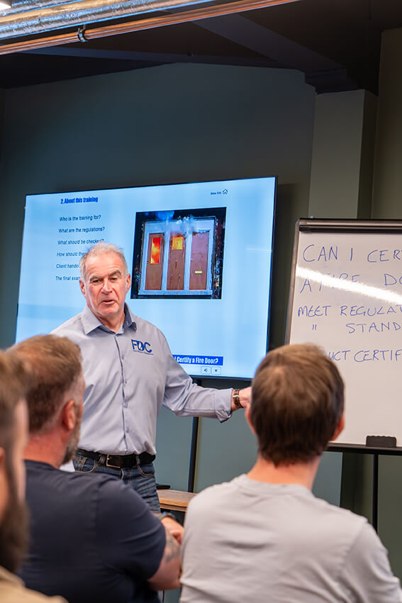 An instructor from Fire Doors Complete leads a "Can I Certify a Fire Door?" training session, with attendees listening and a presentation displayed on the screen.