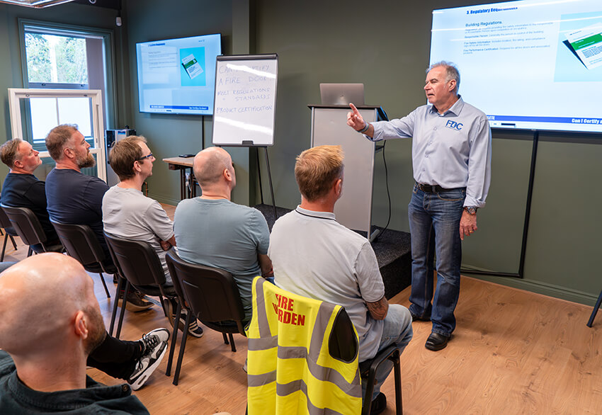 Instructor conducting a "Can I Certify a Fire Door?" training session with attendees seated in a classroom setting.